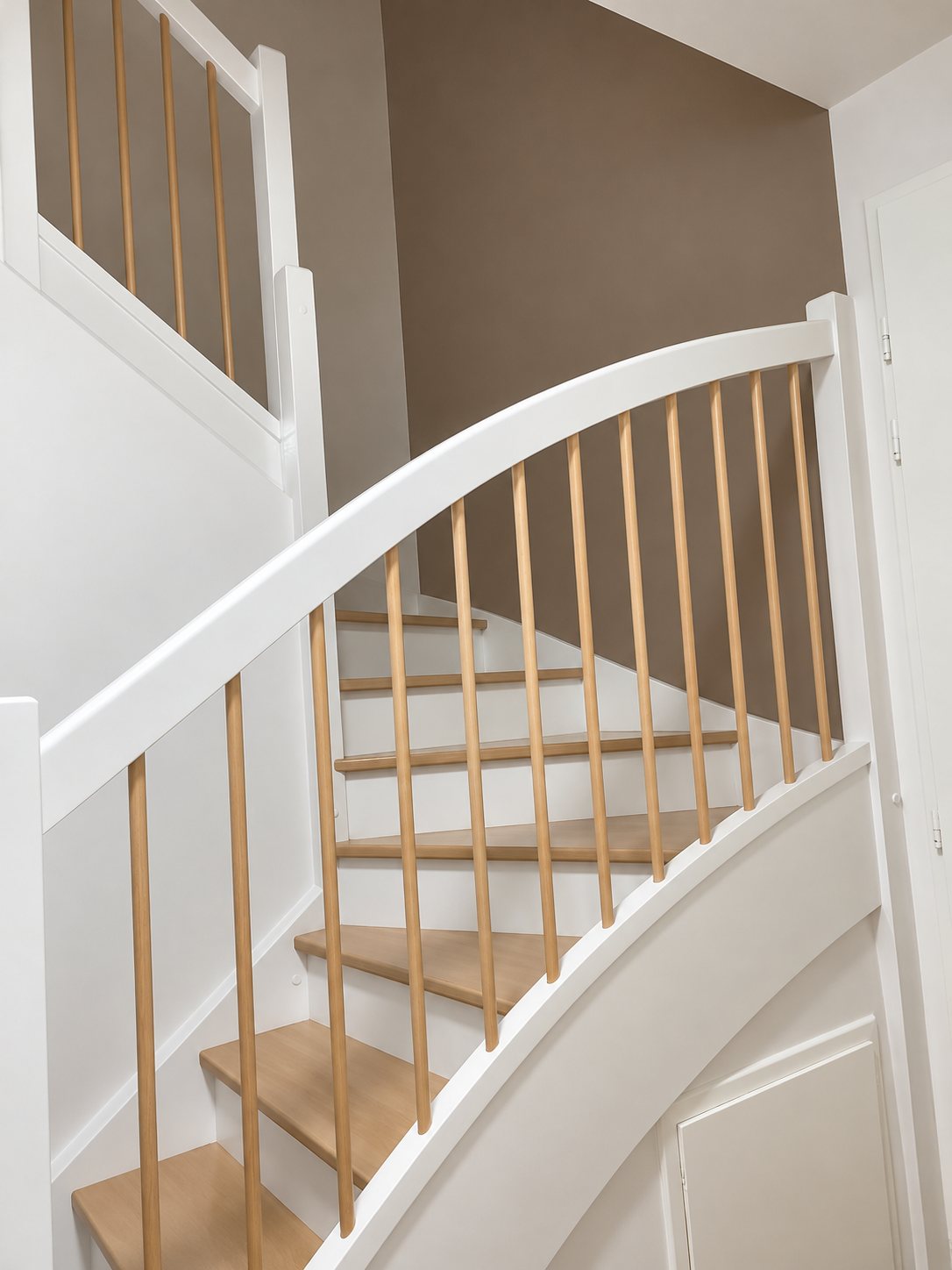Un escalier tournant à la rampe blanche courbe, avec des balustres et des marches en bois clair, dans une cage d'escalier aux murs blancs et taupe.