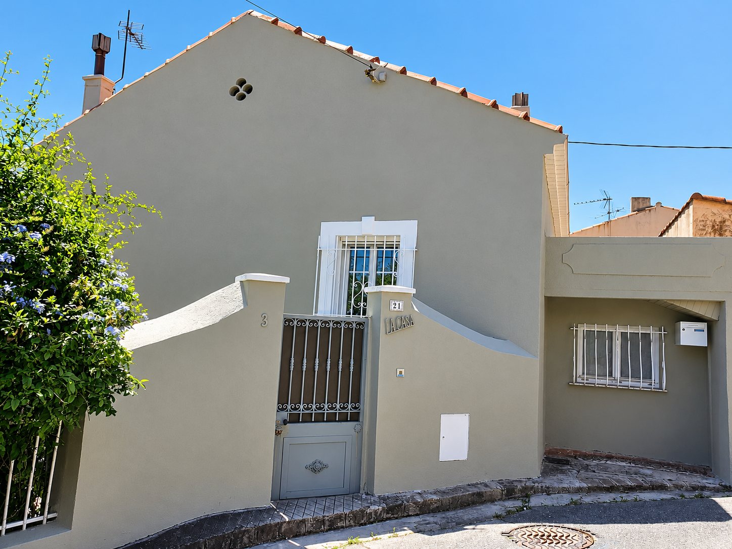 Façade d'une maison aux murs gris-vert avec un portail en fer forgé, une fenêtre grillagée et le nom « La Casa » sur le pilier, sous un ciel bleu.