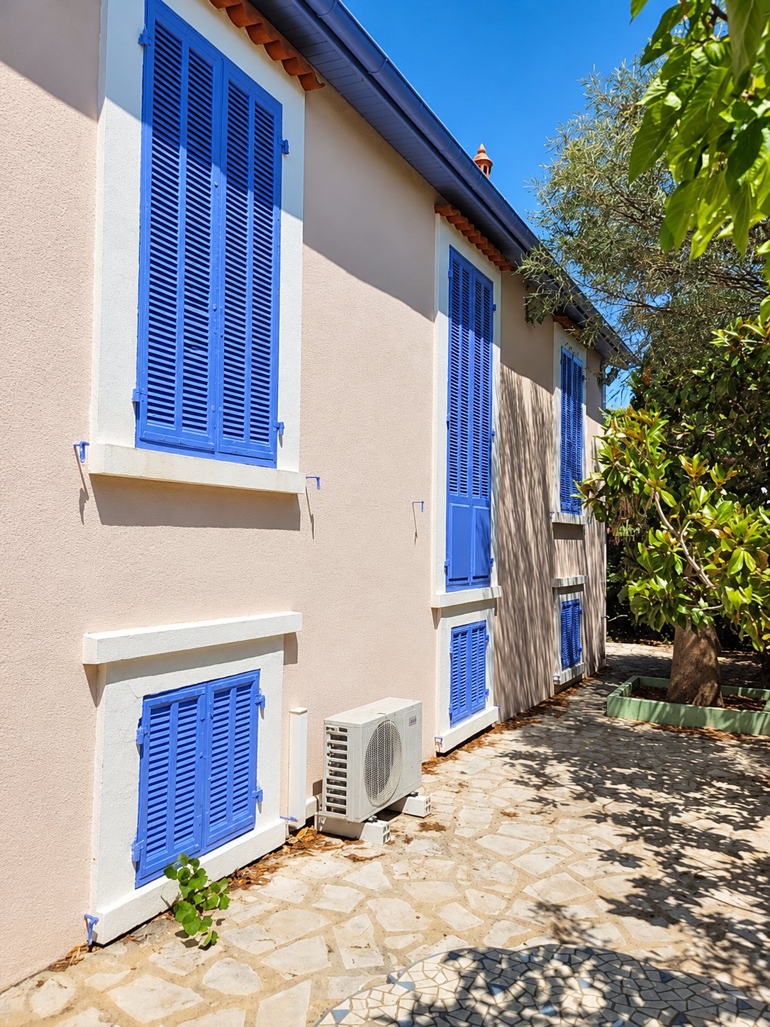 Façade ensoleillée d'une maison aux murs roses et aux volets bleus, avec une unité extérieure de climatisation posée sur un sol dallé de pierres.