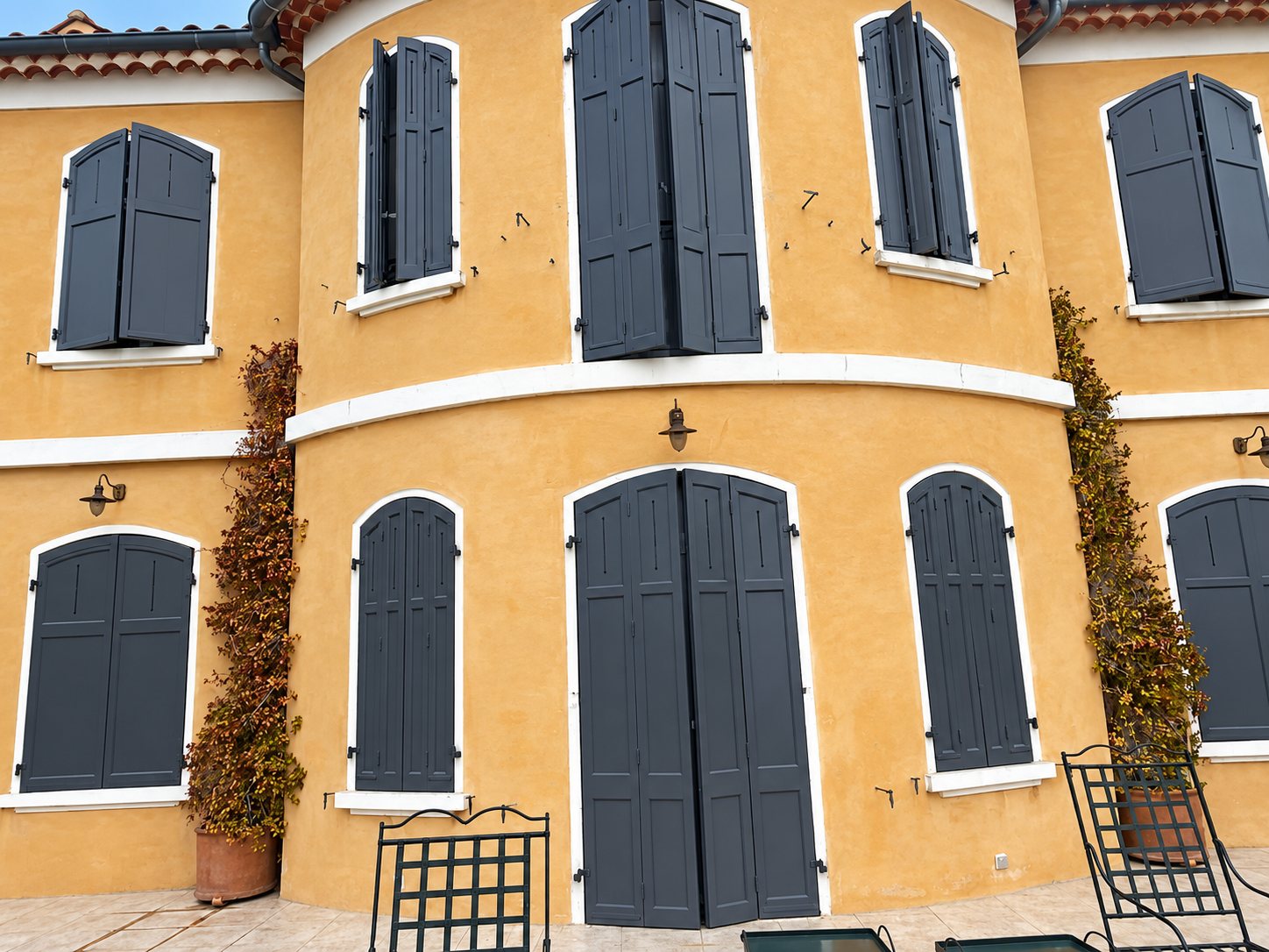 Façade arrondie d'une maison ocre aux encadrements blancs, équipée de volets battants gris anthracite, avec des chaises de jardin en fer au premier plan.