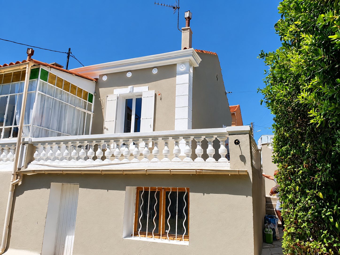 Façade d'une maison aux murs beige clair fraîchement rénovés, avec balustrade blanche, volets blancs, véranda vitrée et fenêtre grillagée sous un ciel bleu.