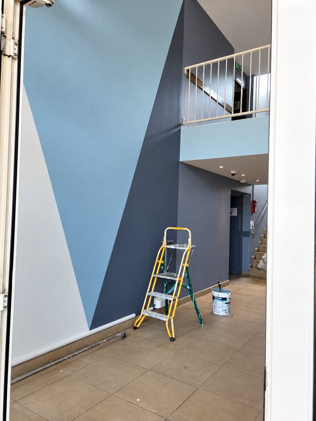 Un escabeau jaune et un pot de peinture sont posés dans un hall d'immeuble dont le mur est peint de grands triangles bleus et gris.