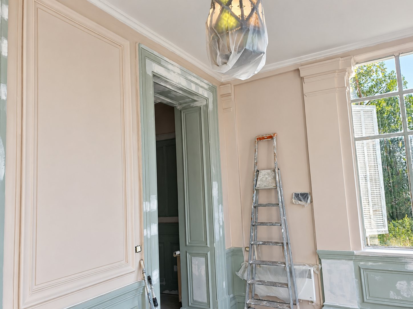 Pièce ancienne en cours de peinture avec boiseries rose pâle et gris-vert, une échelle de peintre, un lustre protégé par une housse plastique et une grande fenêtre.