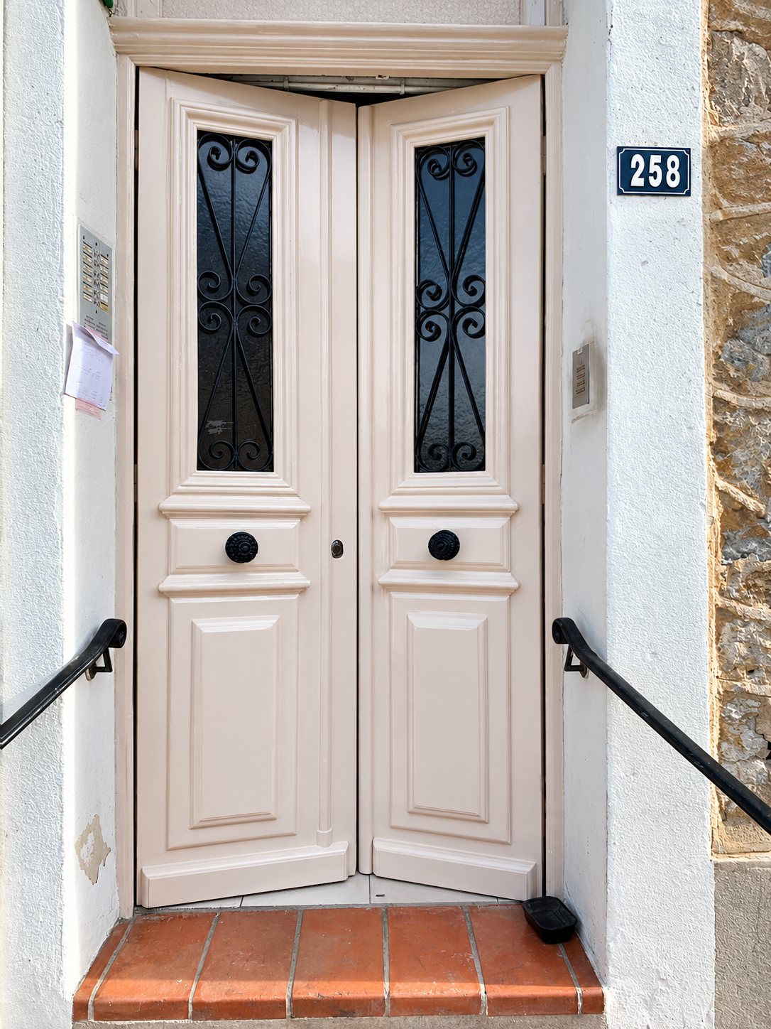 Porte d'entrée à deux battants de couleur rose pâle, ornée de grilles en fer forgé noir, avec le numéro 258 sur le mur et une rampe noire de chaque côté.