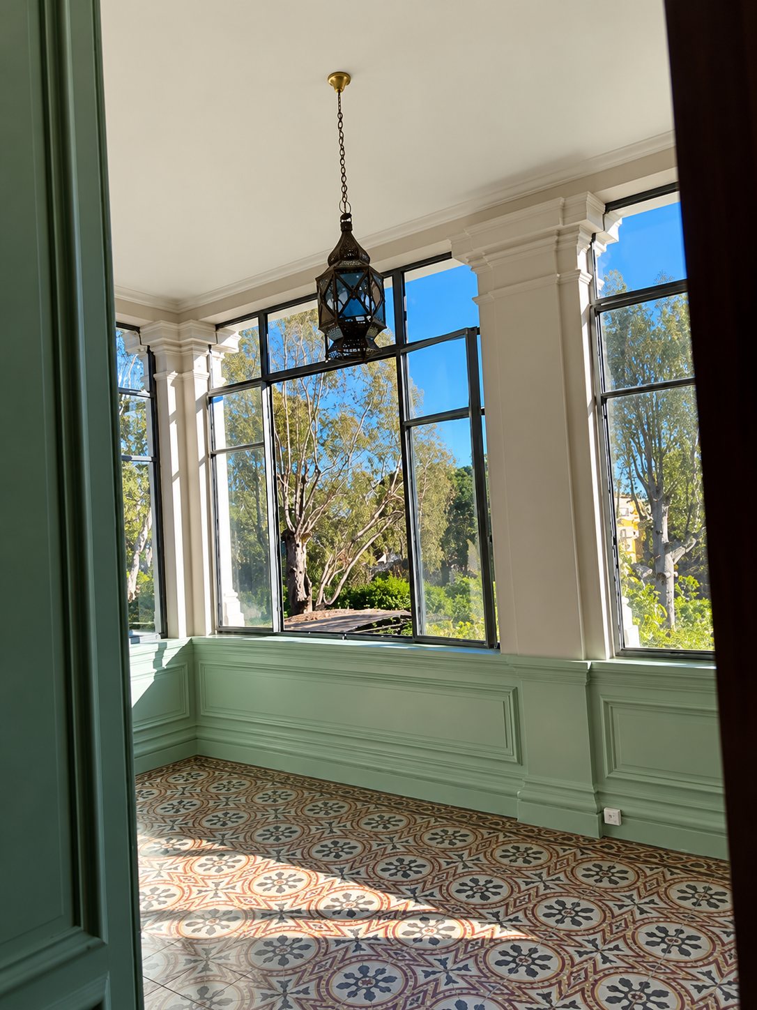 Une véranda aux grandes fenêtres donnant sur des arbres, avec des boiseries vert pâle, un sol en carreaux de ciment décorés et une lanterne ancienne suspendue au plafond.